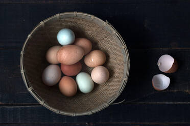 From above shot of wicker bowl filled with raw eggs on wooden table with eggshell near - ADSF38306