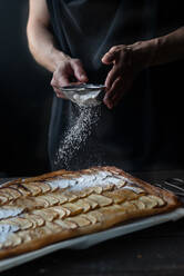 Ausschnittaufnahme einer Frau, die frisch gebackenen Frangipane-Apfelkuchen mit Puderzucker auf einem Holztisch bedeckt - ADSF38310