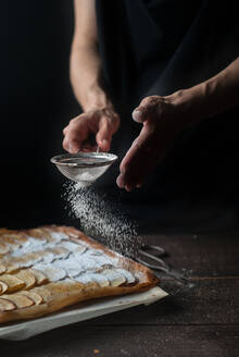 Ausschnittaufnahme einer Frau, die frisch gebackenen Frangipane-Apfelkuchen mit Puderzucker auf einem Holztisch bedeckt - ADSF38311