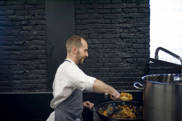 Junger Mann in grauer Schürze rührt in einer großen Metallpfanne in einer Restaurantküche - ADSF38317