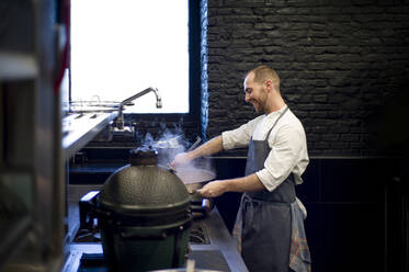 Junger fröhlicher Mann in grauer Schürze rührt in einer großen Metallpfanne in einer Restaurantküche - ADSF38318