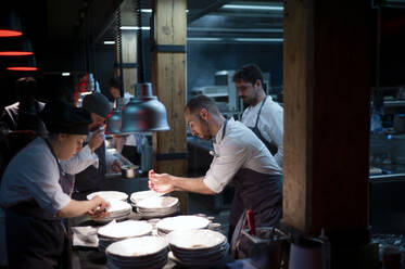 Young cooks men in aprons carefully serving meal in white ceramic dishes on restaurant kitchen - ADSF38324