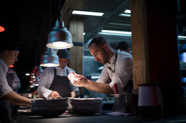 Young cooks men in aprons carefully serving meal in white ceramic dishes on restaurant kitchen - ADSF38325