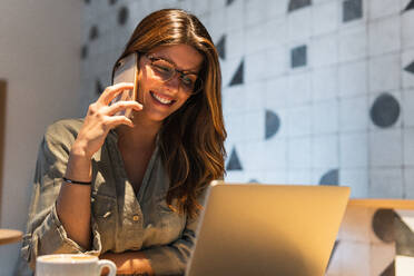 Young happy female in eyewear talking on cellphone while at cafe table with netbook - ADSF38343