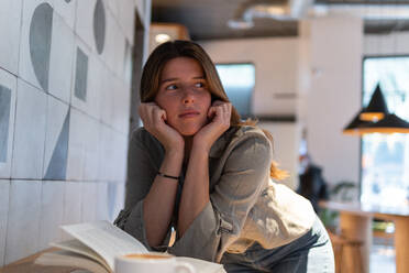 Young wistful female in casual apparel with textbook and cup of hot drink looking away in cafeteria - ADSF38344