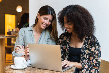 Cheerful multiethnic female friends surfing internet on netbook at table with cup of coffee in cafe - ADSF38346