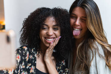 Crop cheerful woman with tongue out near ethnic friend touching face while taking self portrait on cellphone - ADSF38352