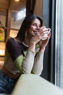 Young content female with cup of hot drink leaning on hand and looking away against window in cafeteria - ADSF38358