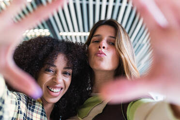 Cheerful ethnic woman near best friend with pouting lips showing focus gesture while looking at camera - ADSF38365