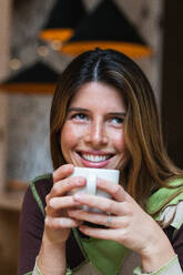 Young dreamy smiling female blowing on hot drink in cup while looking away in cafeteria on blurred background - ADSF38373
