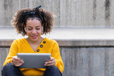 Smiling African American female in wireless earphones sitting on stairs in city and watching movie on tablet - ADSF38376