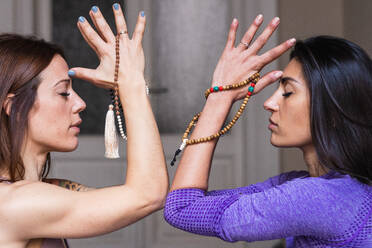 Seitenansicht von friedlichen jungen ethnischen Damen mit Jamala-Gebetsperlen, die mit geschlossenen Augen meditieren, während sie gemeinsam Yoga am Morgen praktizieren - ADSF38388