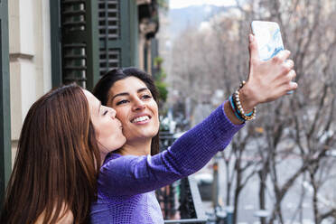 Seitenansicht einer glücklichen jungen hispanischen homosexuellen Frau, die die Wange ihrer lächelnden Freundin küsst, während sie ein Selfie mit ihrem Smartphone auf dem Balkon macht - ADSF38389