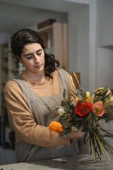 Adult woman in casual clothes and gray apron making beautiful flower composition with dry green plants and fresh flowers while standing at table at home - ADSF38440