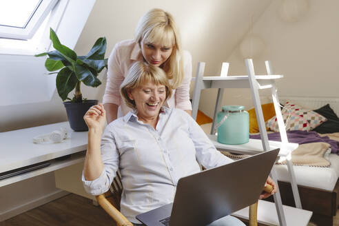 Blonde Frau mit Mutter, die einen Laptop benutzt und auf einem Stuhl im Schlafzimmer sitzt - IHF01278