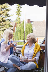 Smiling mother and daughter with coffee cups sitting together in balcony - IHF01286