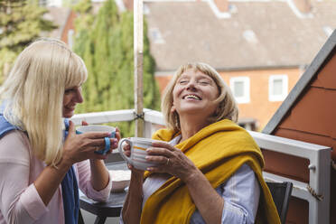 Happy mother and daughter with coffee cups sitting together in balcony - IHF01287