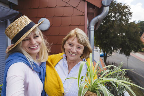 Happy senior woman with daughter wearing hat standing in balcony - IHF01292