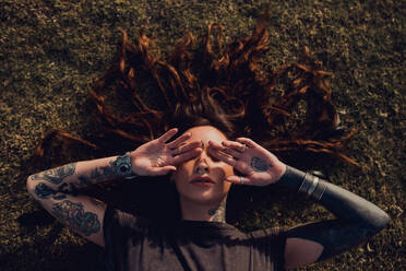 From above serene stylish brunette in casual clothes with tattoos and piercing laying on grassy glade and covering face with hands while relaxing alone in park - ADSF38459