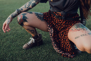 Von oben unerkennbar hockende Frau mit Tattoos in schwarzem Hemd und trendigem bunt gepunktetem Rock in Komposition mit Lederschuhen auf grüner Wiese im Park - ADSF38470