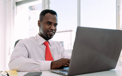Smiling black male entrepreneur working on netbook in modern office - ADSF38530