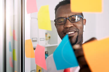 Blick durch eine Glaswand auf einen zufriedenen afroamerikanischen männlichen Unternehmer mit Brille und bunten Papieraufklebern im Büro - ADSF38534