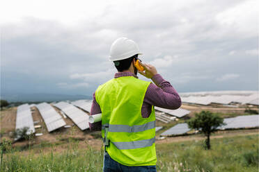 Rückenansicht eines nicht erkennbaren Mannes, der eine Blaupause hält, während er mit einem Walkie-Talkie vor einem Solarkraftwerk und einem bedeckten grauen Himmel spricht - ADSF38545