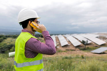 Seitenansicht eines Mannes, der einen Bauplan hält, während er mit einem Walkie-Talkie vor einem Solarkraftwerk und einem bedeckten grauen Himmel spricht - ADSF38549