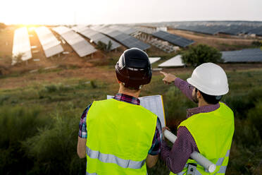 Rückenansicht von anonymen männlichen Kollegen, die an einem bedeckten Tag auf der Baustelle den Bau eines Solarkraftwerks besprechen - ADSF38555