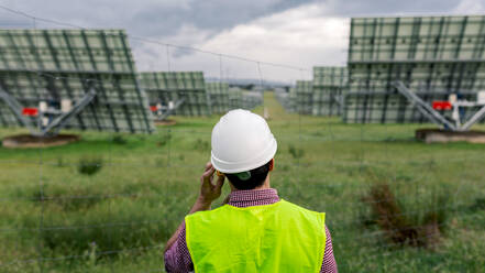 Rückenansicht eines nicht erkennbaren Mannes, der mit einem Walkie-Talkie vor einem Solarkraftwerk und einem bedeckten grauen Himmel spricht - ADSF38556