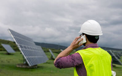 Back view of anonymous man in hardhat at photovoltaic panels while discussing work of solar power station on smartphone - ADSF38557