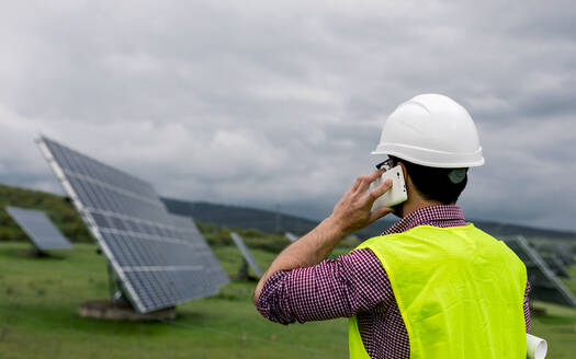 Rückenansicht eines anonymen Mannes mit Schutzhelm an Photovoltaik-Paneelen, während er die Arbeit eines Solarkraftwerks auf einem Smartphone diskutiert - ADSF38557