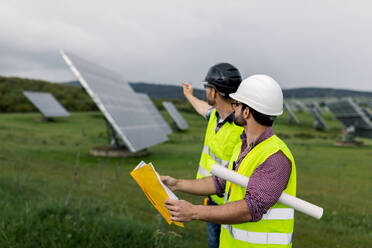 Seitenansicht von männlichen Kollegen, die den Bau eines Solarkraftwerks an einem bedeckten Tag auf der Baustelle besprechen - ADSF38558