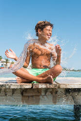 Positiver Jugendlicher, der auf einem hölzernen Kai sitzt und an einem sonnigen Sommertag im Wasser plantscht - ADSF38609