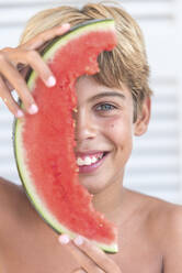 Fröhlicher Teenager, der sein Gesicht mit einem Stück saftiger Wassermelone bedeckt, während er in die Kamera schaut und das Sommerwochenende genießt - ADSF38612