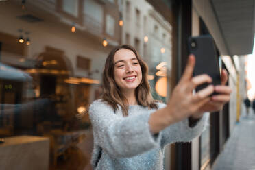 Attraktive lächelnde Frau, die in der Nähe eines Cafés steht und ein Selfie mit ihrem Smartphone macht, während sie das Wochenende in der Stadt genießt - ADSF38628