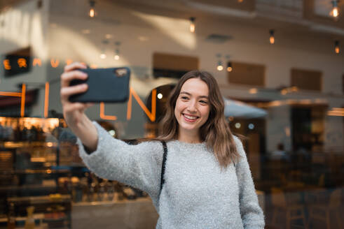 Attraktive lächelnde Frau, die in der Nähe eines Cafés steht und ein Selfie mit ihrem Smartphone macht, während sie das Wochenende in der Stadt genießt - ADSF38629