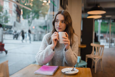 Ruhige Frau, die mit einer Tasse Kaffee und einem Notizbuch am Tisch sitzt und das Wochenende im Café genießt, während sie die Straße beobachtet und aus dem Fenster schaut - ADSF38631