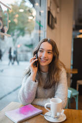 Inhalt Charmante Frau sitzt am Tisch im Café und spricht mit ihrem Smartphone, während sie aus dem Fenster schaut und lächelt - ADSF38632