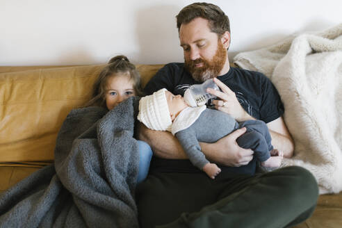 Mädchen (2-3), das dem Vater beim Füttern des neugeborenen Sohnes (0-1 Monate) auf dem Sofa hilft - TETF01759