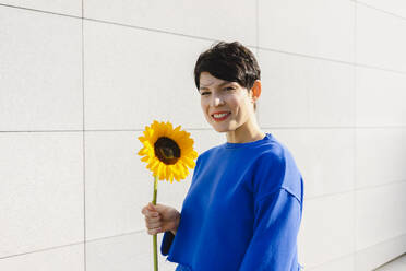 Glückliche Frau mit Sonnenblume an einer Mauer stehend an einem sonnigen Tag - SEAF01198