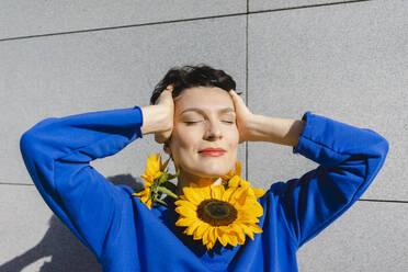 Lächelnde Frau mit Sonnenblumen um den Hals vor einer Mauer stehend an einem sonnigen Tag - SEAF01205