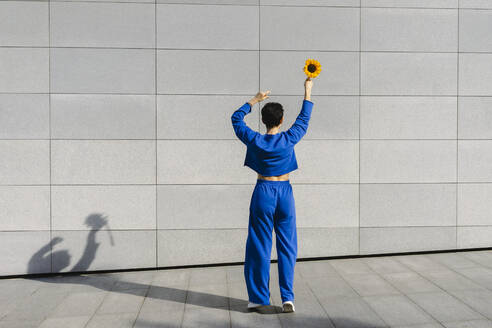 Frau mit erhobenen Armen, die eine Sonnenblume vor einer Mauer hält, an einem sonnigen Tag - SEAF01209