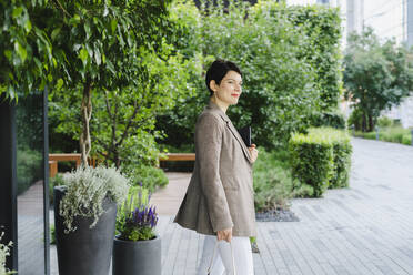 Smiling businesswoman with tablet PC standing by plants at office park - SEAF01215