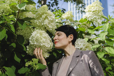 Lächelnde Geschäftsfrau, die an Blumen riecht, neben einer Pflanze stehend - SEAF01224