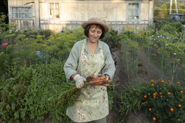 Glückliche ältere Frau mit frisch gepflückten Möhren im Garten stehend - VBUF00178