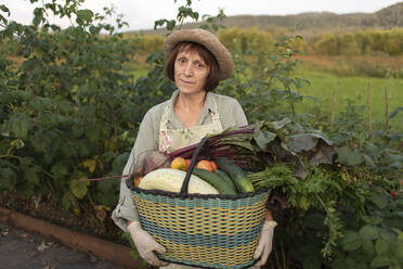 Lächelnde ältere Frau mit einem Korb mit frisch geerntetem Gemüse an einer Pflanze stehend - VBUF00185