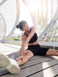 Smiling woman practicing stretching exercise sitting on footpath - AMRF00010