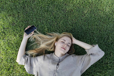 Woman with eyes closed holding mobile phone listening music through wireless in-ear headphones lying on grass - TYF00414