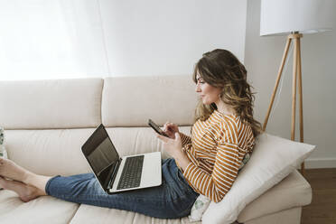 Junge Frau mit Laptop und Mobiltelefon auf dem Sofa - EBBF06402